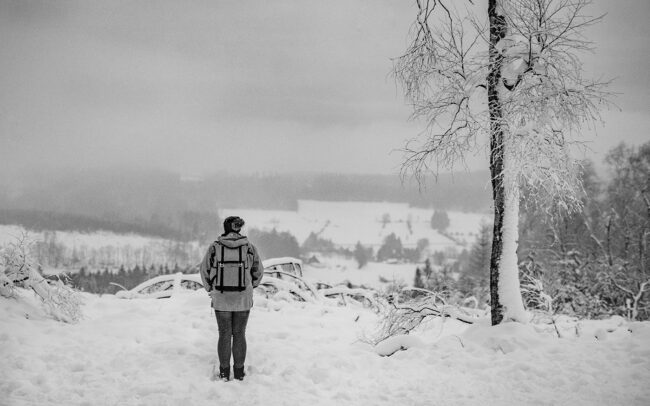 Eine Fotografin mit Wintermütze und Rucksack steht mit dem Rücken zur Kamera und blickt auf die nebelige Schneelandschaft