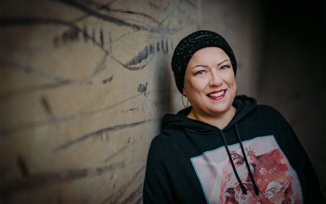 Fotografin mit Wintermütze und Hoodiekleid steht lächelnd an einer Wand angelehnt