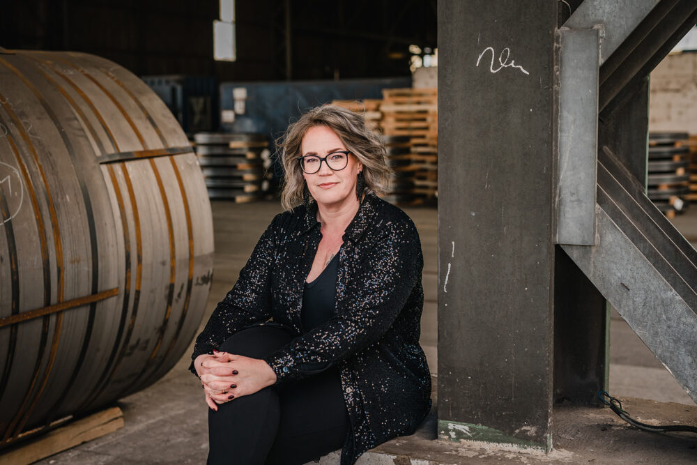 Portrait einer Frau, die in einer Industrielagerhalle neben einer Stahlsäule sitzt und in die Kamera lächelt