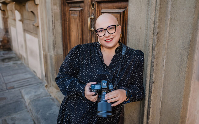 Fotografin im gepunkteten Kleid lehnt lächelnd an einer Mauer in Florenz und hat ihre Kamera in der Hand